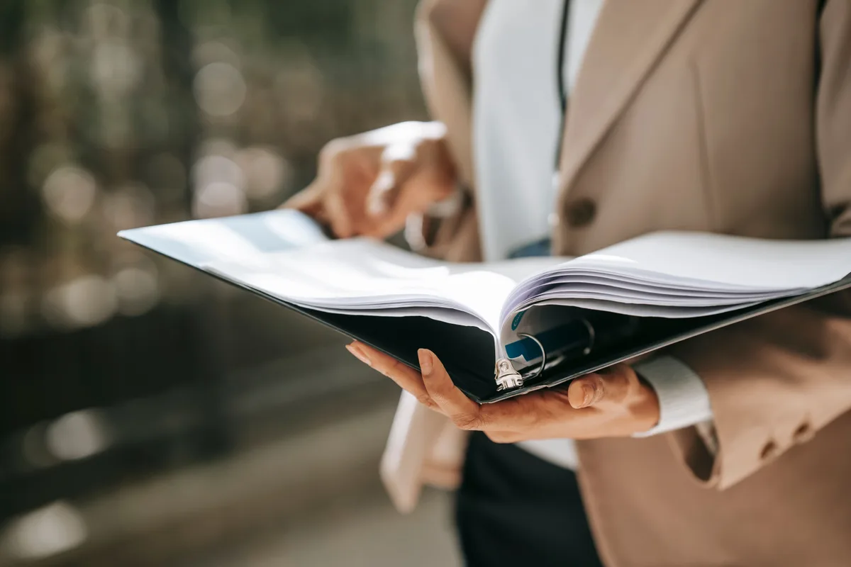 Une professionnelle en veste beige feuillette les pages d'un classeur ouvert, tenu à deux mains