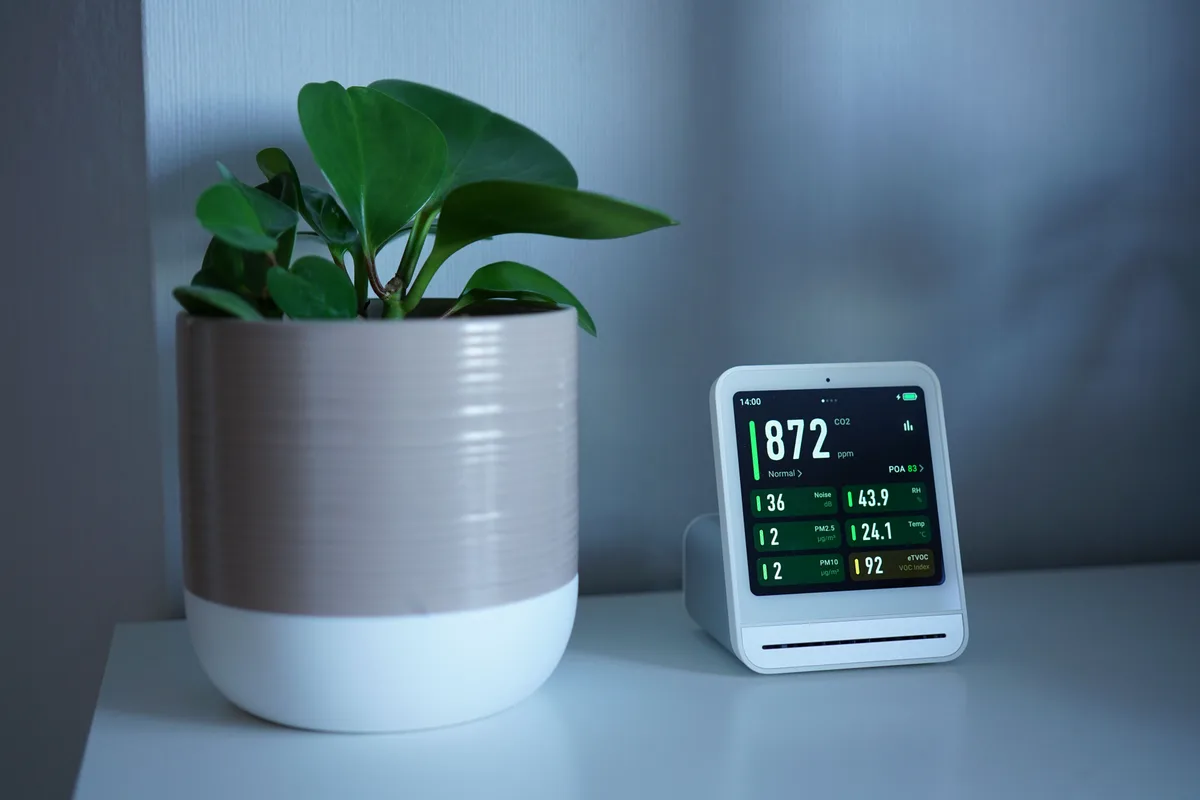 Capteur de qualité de l'air affichant un taux de CO2 de 872 ppm, posé à côté d'une plante verte
