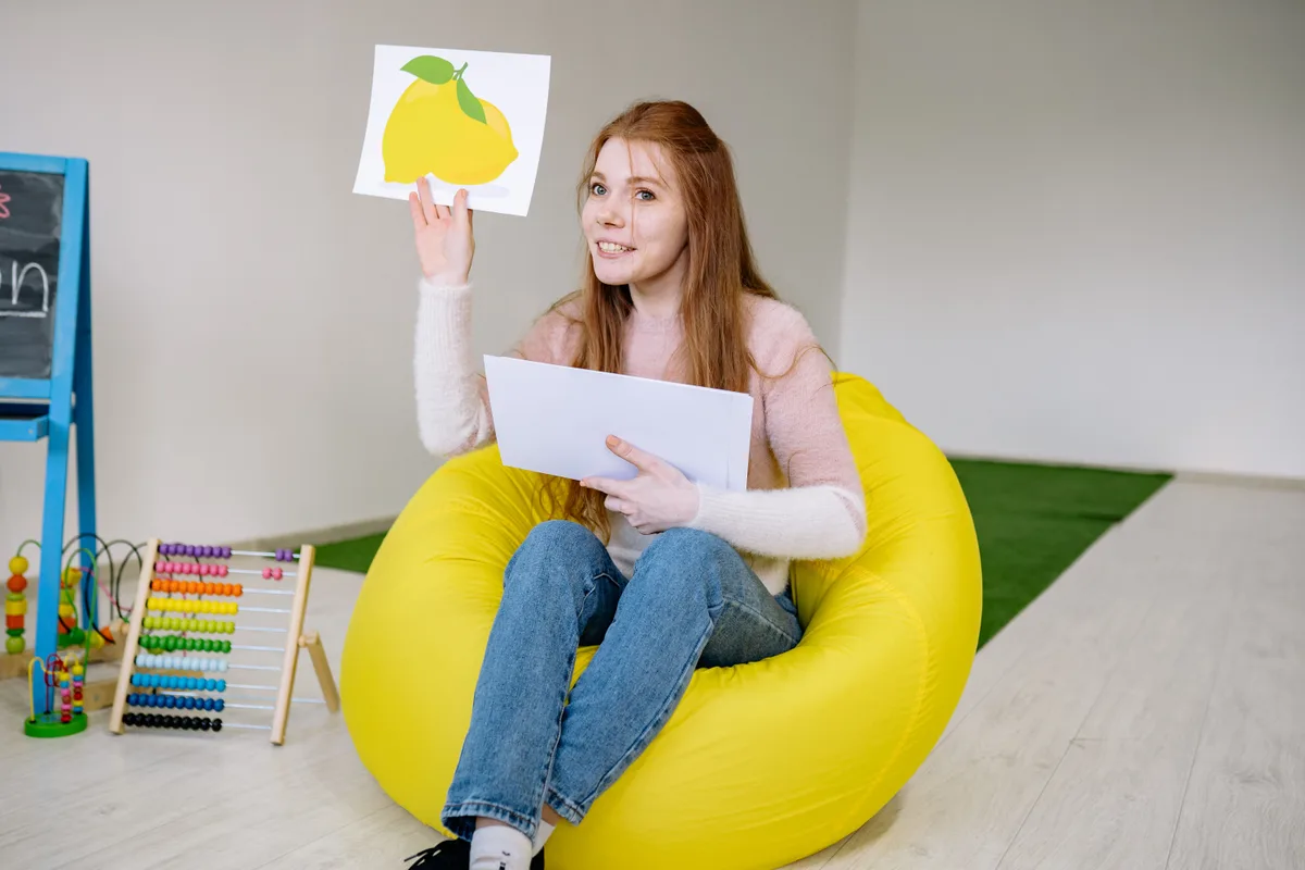 Une professionnelle de la petite enfance souriante, assise sur un pouf jaune dans une salle d'activités, tient une carte illustrée à la main