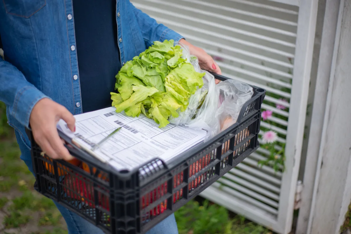Personne portant une cagette de légumes frais avec les bons de livraison posés dessus